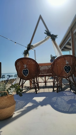 A beachside setting with two wicker chairs facing a decorated triangular arch draped with white fabric and greenery. The scene is sunlit, with shadows cast on a wooden deck. A basket with plants is placed in the foreground, adding to the coastal ambiance.