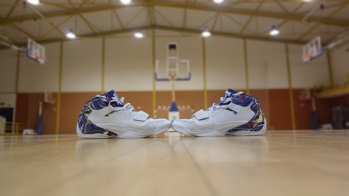 A pair of white athletic shoes with colorful details is positioned on the floor of an indoor basketball court. The court features wooden flooring, and two basketball hoops are visible in the background. The setting is well-lit with overhead lights and has a high ceiling with exposed beams.