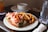 A plate holds a lobster roll filled with lobster meat and lettuce alongside a serving of golden brown French fries. In the background, a white bowl on a saucer and a tall glass of water is visible. The setting appears to be a casual dining table with soft natural lighting.