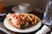 A plate holds a lobster roll filled with lobster meat and lettuce alongside a serving of golden brown French fries. In the background, a white bowl on a saucer and a tall glass of water is visible. The setting appears to be a casual dining table with soft natural lighting.