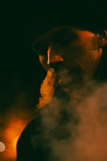 A dimly lit portrait captures a person exhaling smoke, which is illuminated by warm, orange lighting. The face is partially obscured by shadows and the wispy trails of smoke, creating a mysterious and atmospheric scene.