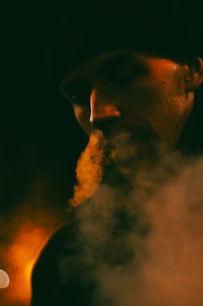 A dimly lit portrait captures a person exhaling smoke, which is illuminated by warm, orange lighting. The face is partially obscured by shadows and the wispy trails of smoke, creating a mysterious and atmospheric scene.