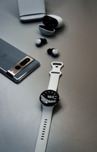 A minimalistic setup featuring a gray smartphone, black and white wireless earbuds with their charging case, and a white wristwatch with a digital display, all placed on a smooth, neutral gray surface.