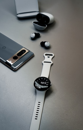 A minimalistic setup featuring a gray smartphone, black and white wireless earbuds with their charging case, and a white wristwatch with a digital display, all placed on a smooth, neutral gray surface.