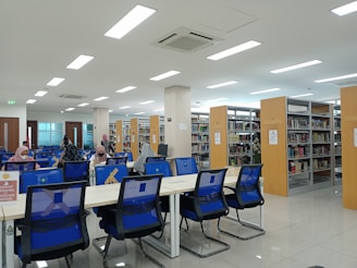 A spacious, well-lit library with rows of bookshelves and several tables with blue chairs. People are seated at the tables, engaged in reading or working on desks. The interior features large overhead lights and a clean, modern design. Signage is visible, encouraging quiet behavior.