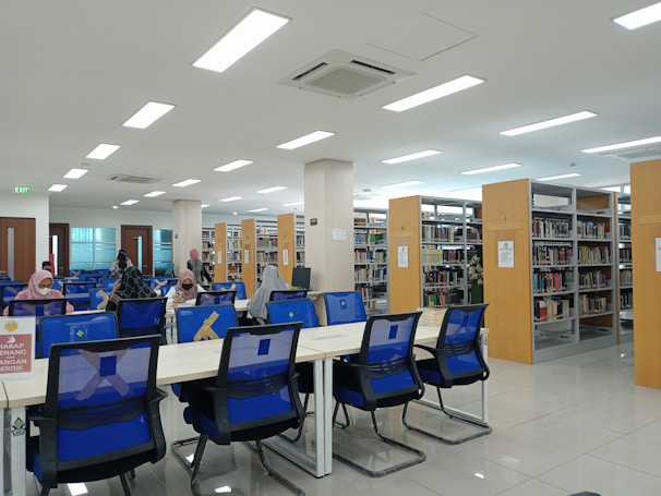 A spacious, well-lit library with rows of bookshelves and several tables with blue chairs. People are seated at the tables, engaged in reading or working on desks. The interior features large overhead lights and a clean, modern design. Signage is visible, encouraging quiet behavior.