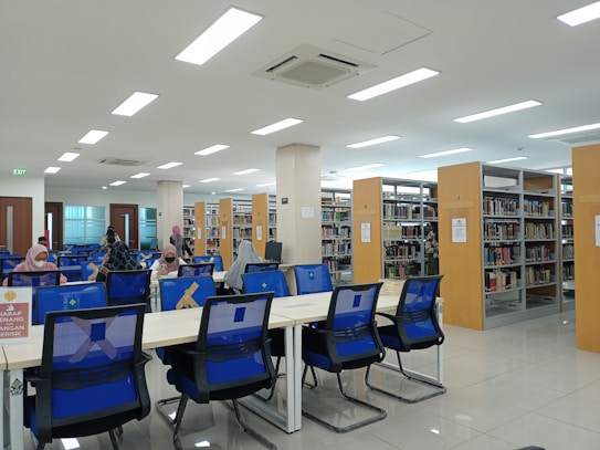 A spacious, well-lit library with rows of bookshelves and several tables with blue chairs. People are seated at the tables, engaged in reading or working on desks. The interior features large overhead lights and a clean, modern design. Signage is visible, encouraging quiet behavior.