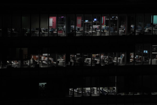 The image depicts a dimly lit office building at night, where several floors are visible through the windows. Each window reveals individual office spaces with computers, desks, and chairs. There are only a few lights illuminated within these offices, suggesting late-night work or an empty building.