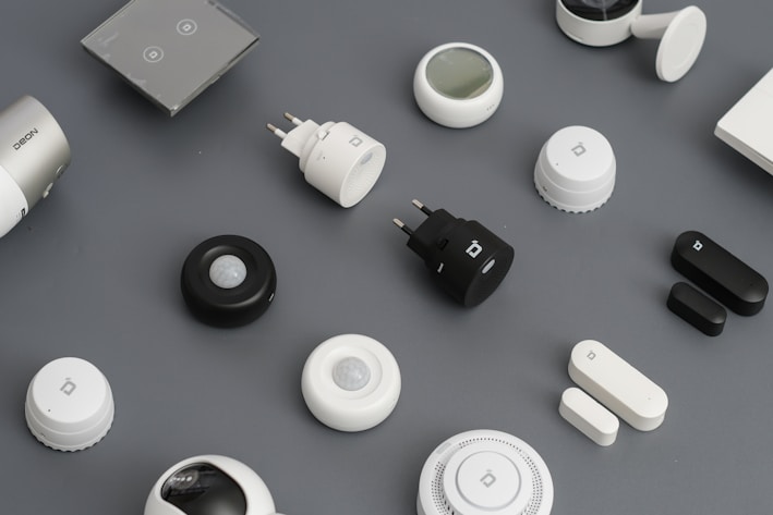 A variety of smart home devices are arranged on a flat surface, including sensors, plugs, and hubs. The devices are mostly black and white, with some showing LED indicators and branding. The background is a muted gray, emphasizing the sleek and modern design of the gadgets.