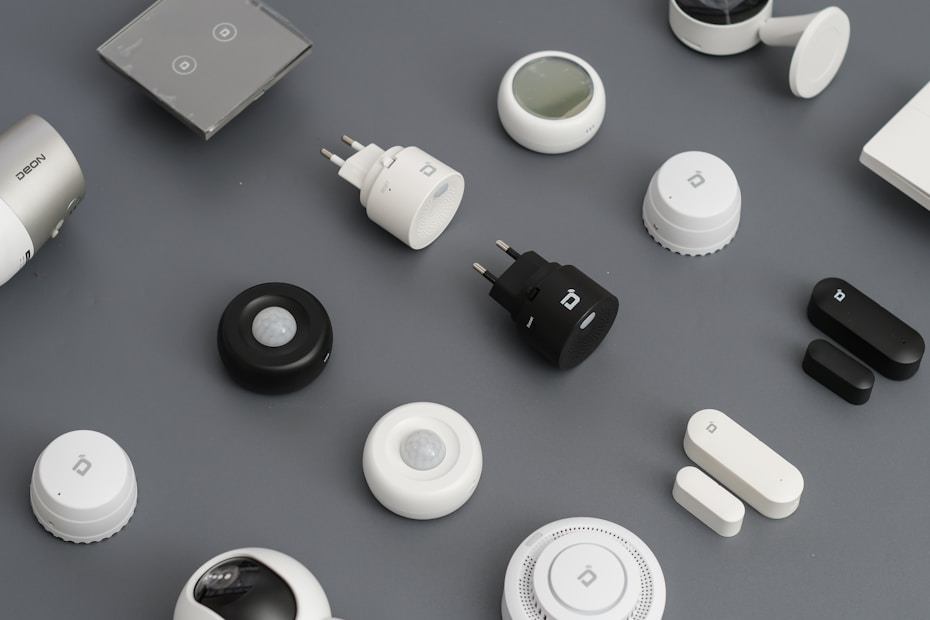 A variety of smart home devices are arranged on a flat surface, including sensors, plugs, and hubs. The devices are mostly black and white, with some showing LED indicators and branding. The background is a muted gray, emphasizing the sleek and modern design of the gadgets.