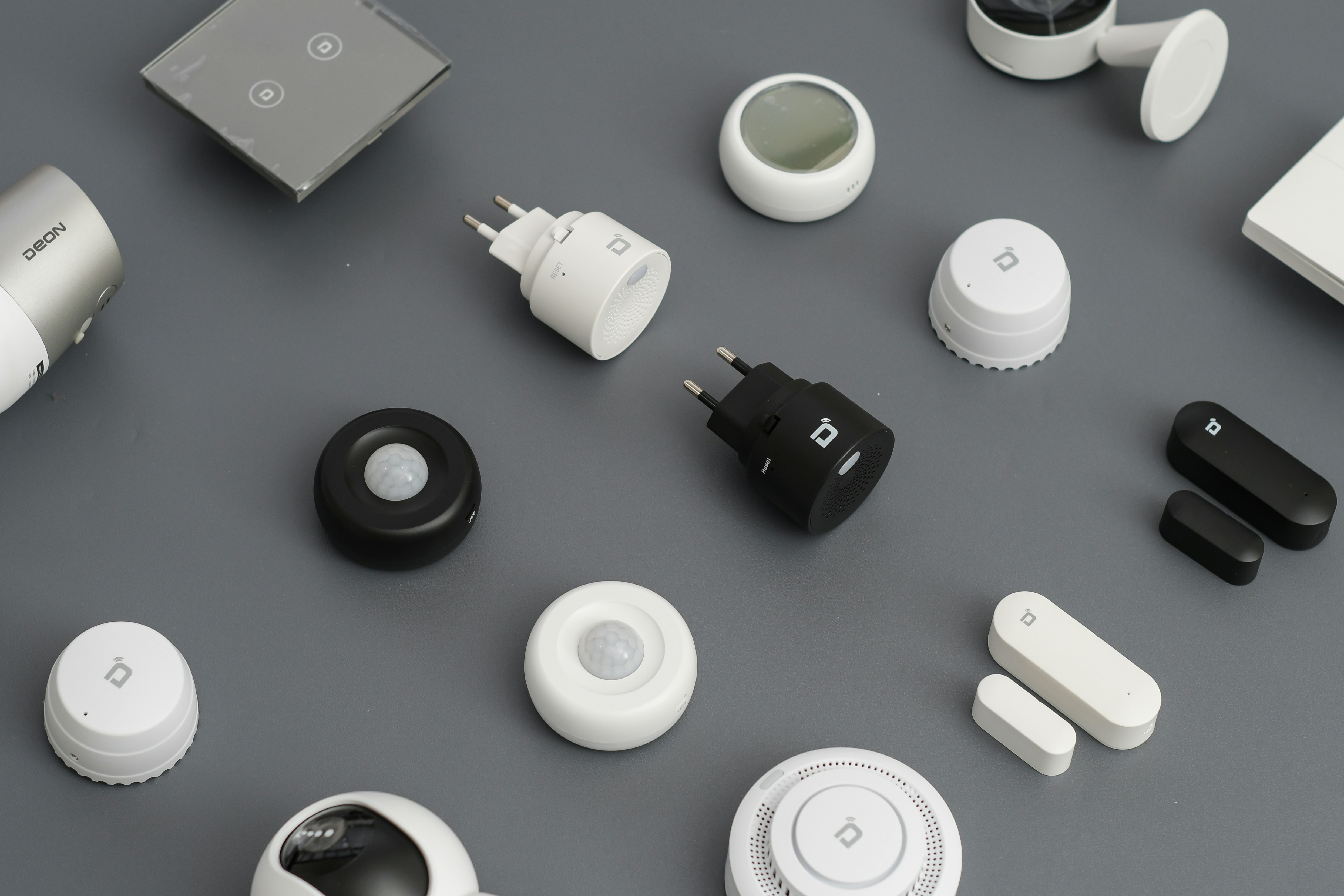 A variety of smart home devices are arranged on a flat surface, including sensors, plugs, and hubs. The devices are mostly black and white, with some showing LED indicators and branding. The background is a muted gray, emphasizing the sleek and modern design of the gadgets.