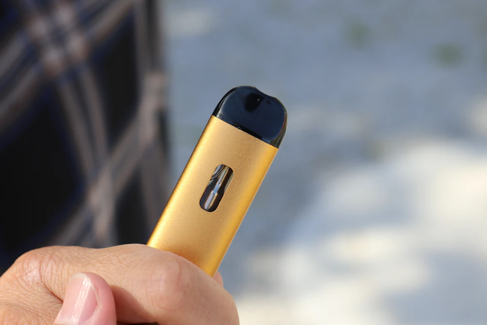 The image shows a close-up view of a person's hand holding an electronic cigarette, or e-cigarette, with a gold finish and black top. The e-cigarette is positioned on the right side of the frame, with its sleek design and smooth surface reflecting light. In the blurred background, there are trees visible, suggesting that the scene takes place outdoors.