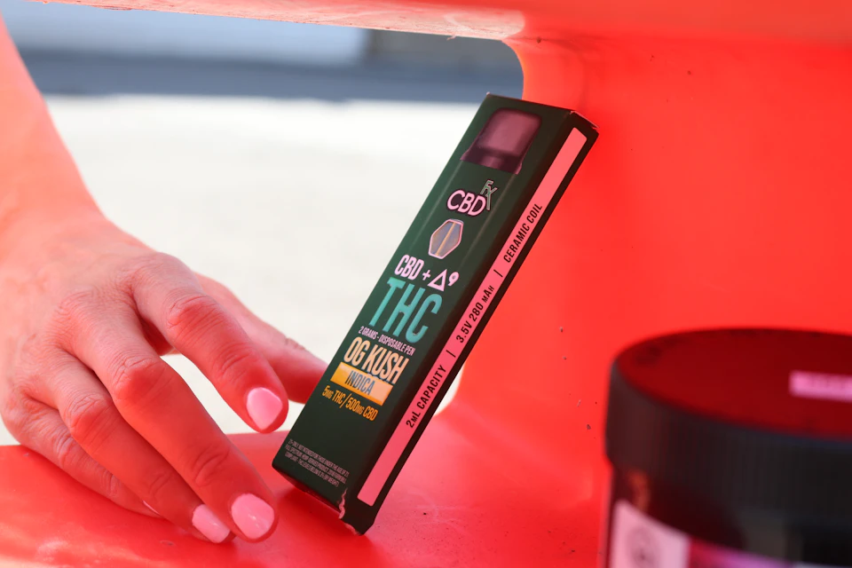 The image shows a person's hand reaching out towards an open green box containing what appears to be a joint or marijuana product, likely THC edibles. The box is resting on a red shelf against a white wall. In the background, there are two other objects - one black and one brown.