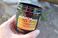 A hand is holding a dark-colored container labeled 'Reserve CBD/THC Full Spectrum Hemp Extract' with details about the product's dosage and type. The container mentions 30 gummies and a flavor of peach. The background is blurred, possibly showing an outdoor setting.