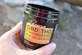 A hand is holding a dark-colored container labeled 'Reserve CBD/THC Full Spectrum Hemp Extract' with details about the product's dosage and type. The container mentions 30 gummies and a flavor of peach. The background is blurred, possibly showing an outdoor setting.