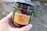 A hand is holding a dark-colored container labeled 'Reserve CBD/THC Full Spectrum Hemp Extract' with details about the product's dosage and type. The container mentions 30 gummies and a flavor of peach. The background is blurred, possibly showing an outdoor setting.