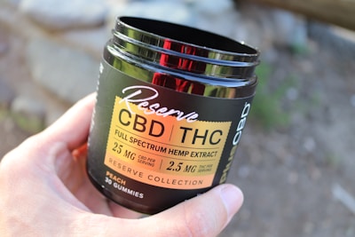 A hand is holding a dark-colored container labeled 'Reserve CBD/THC Full Spectrum Hemp Extract' with details about the product's dosage and type. The container mentions 30 gummies and a flavor of peach. The background is blurred, possibly showing an outdoor setting.