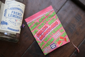 A large glass bottle labeled 'French Lemonade' is lying on a wooden surface next to a bright pink package of watermelon-flavored gummies. The package includes the words 'Delta Munchies' and indicates that it contains hemp-derived Delta 8 THC. Several small, square gummies with a watermelon motif are displayed on the package.