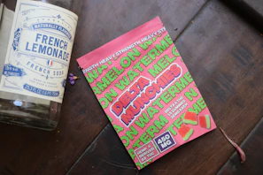 A large glass bottle labeled 'French Lemonade' is lying on a wooden surface next to a bright pink package of watermelon-flavored gummies. The package includes the words 'Delta Munchies' and indicates that it contains hemp-derived Delta 8 THC. Several small, square gummies with a watermelon motif are displayed on the package.