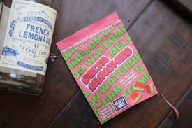 A large glass bottle labeled 'French Lemonade' is lying on a wooden surface next to a bright pink package of watermelon-flavored gummies. The package includes the words 'Delta Munchies' and indicates that it contains hemp-derived Delta 8 THC. Several small, square gummies with a watermelon motif are displayed on the package.