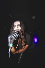 A person with dreadlocks and face paint is holding a microphone while performing. They are wearing black gloves and the atmosphere is dimly lit with some colored lights visible in the background.