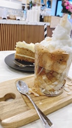 A frothy iced beverage topped with whipped cream and a striped straw, served on a wooden board with a spoon. In the background, a slice of layered cake with cream frosting is placed on a black plate. The setting appears to be a cozy cafe with wooden decor and flowers.