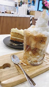 A frothy iced beverage topped with whipped cream and a striped straw, served on a wooden board with a spoon. In the background, a slice of layered cake with cream frosting is placed on a black plate. The setting appears to be a cozy cafe with wooden decor and flowers.