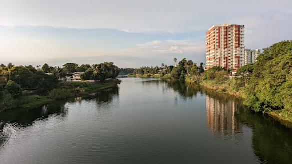 A serene river flows gently with lush green trees lining its banks. A tall residential building stands prominently on the right, reflecting on the water's surface. The sky is clear with a few clouds, indicating a peaceful day.