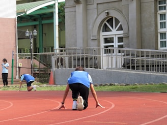 Two people are preparing to start a race on a curved, red athletic track in front of a large building. They are wearing blue uniforms. Another person on the left stands near the track, looking at a clipboard. In the background, there is a building with a large arched doorway and a railing.