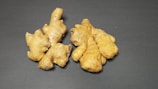 Several pieces of fresh ginger root are placed on a dark, flat surface. The ginger is light brown with a rough, knobby texture, and the lighting highlights its natural curves and intricate shapes.