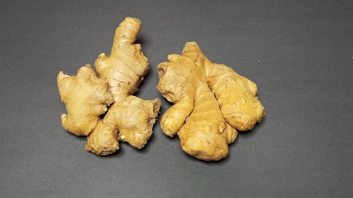 Several pieces of fresh ginger root are placed on a dark, flat surface. The ginger is light brown with a rough, knobby texture, and the lighting highlights its natural curves and intricate shapes.