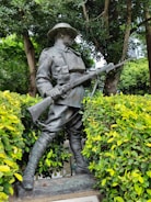 A bronze statue of a soldier in uniform holding a rifle, surrounded by vibrant green foliage and trees. The soldier is depicted in a poised stance, wearing a hat and boots, with a determined expression.