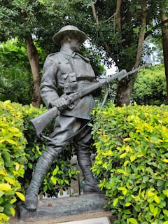 A bronze statue of a soldier in uniform holding a rifle, surrounded by vibrant green foliage and trees. The soldier is depicted in a poised stance, wearing a hat and boots, with a determined expression.