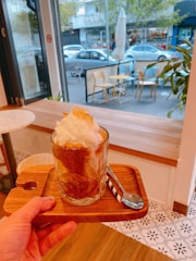 A hand is holding a wooden tray with a glass of coffee topped with whipped cream. The tray includes a spoon with a striped handle. The background shows a cafe with indoor seating, as well as an outdoor view of chairs, tables, and parked cars. The scene appears cozy and inviting.