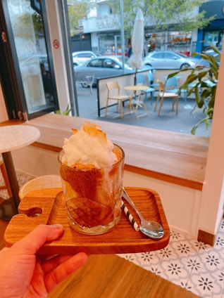 A hand is holding a wooden tray with a glass of coffee topped with whipped cream. The tray includes a spoon with a striped handle. The background shows a cafe with indoor seating, as well as an outdoor view of chairs, tables, and parked cars. The scene appears cozy and inviting.