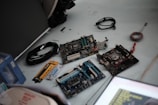 A collection of computer hardware components placed on a light-colored surface. Several computer motherboards are visible along with various cables, a syringe-like thermal paste applicator, and a yellow ruler. Some loose wires and a black cable are also present.