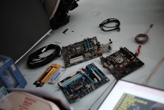 A collection of computer hardware components placed on a light-colored surface. Several computer motherboards are visible along with various cables, a syringe-like thermal paste applicator, and a yellow ruler. Some loose wires and a black cable are also present.