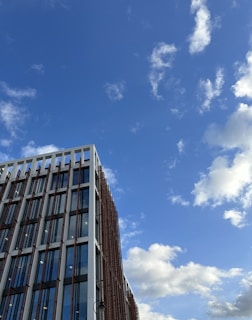 A modern building with large glass windows and a prominent grid-like structure on its facade stands against a backdrop of a bright blue sky with scattered white clouds.