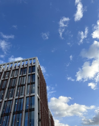 A modern building with large glass windows and a prominent grid-like structure on its facade stands against a backdrop of a bright blue sky with scattered white clouds.