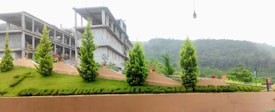 A landscape featuring a series of small, well-maintained shrubs and plants arranged on a grassy slope with carved shapes. In the background, there is a multi-storey concrete building under construction, and the setting is bordered by a lush forested hill.
