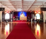 A large indoor space with a polished wooden floor and a red carpet leading to a central altar. The altar is ornately decorated with religious imagery and flanked by two large windows with stained glass. Chairs are arranged in rows, and the ceiling features multiple fans and lights.