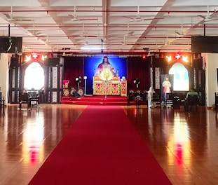 A large indoor space with a polished wooden floor and a red carpet leading to a central altar. The altar is ornately decorated with religious imagery and flanked by two large windows with stained glass. Chairs are arranged in rows, and the ceiling features multiple fans and lights.