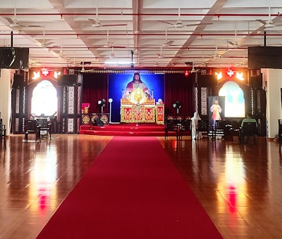A large indoor space with a polished wooden floor and a red carpet leading to a central altar. The altar is ornately decorated with religious imagery and flanked by two large windows with stained glass. Chairs are arranged in rows, and the ceiling features multiple fans and lights.