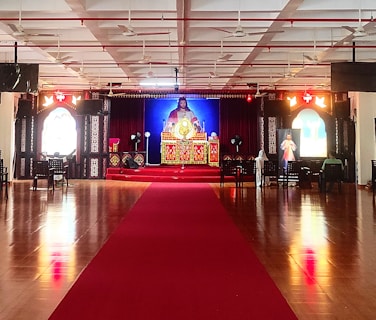 A large indoor space with a polished wooden floor and a red carpet leading to a central altar. The altar is ornately decorated with religious imagery and flanked by two large windows with stained glass. Chairs are arranged in rows, and the ceiling features multiple fans and lights.