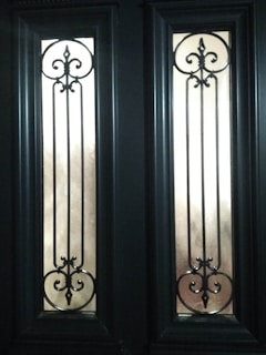 Two tall, narrow glass panels are set within a dark wooden door, each featuring intricate wrought iron scrollwork designs with symmetrical curves and points. The panels allow diffused light to pass through, casting soft shadows.