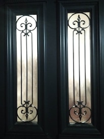 Two tall, narrow glass panels are set within a dark wooden door, each featuring intricate wrought iron scrollwork designs with symmetrical curves and points. The panels allow diffused light to pass through, casting soft shadows.