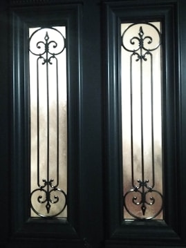 Two tall, narrow glass panels are set within a dark wooden door, each featuring intricate wrought iron scrollwork designs with symmetrical curves and points. The panels allow diffused light to pass through, casting soft shadows.