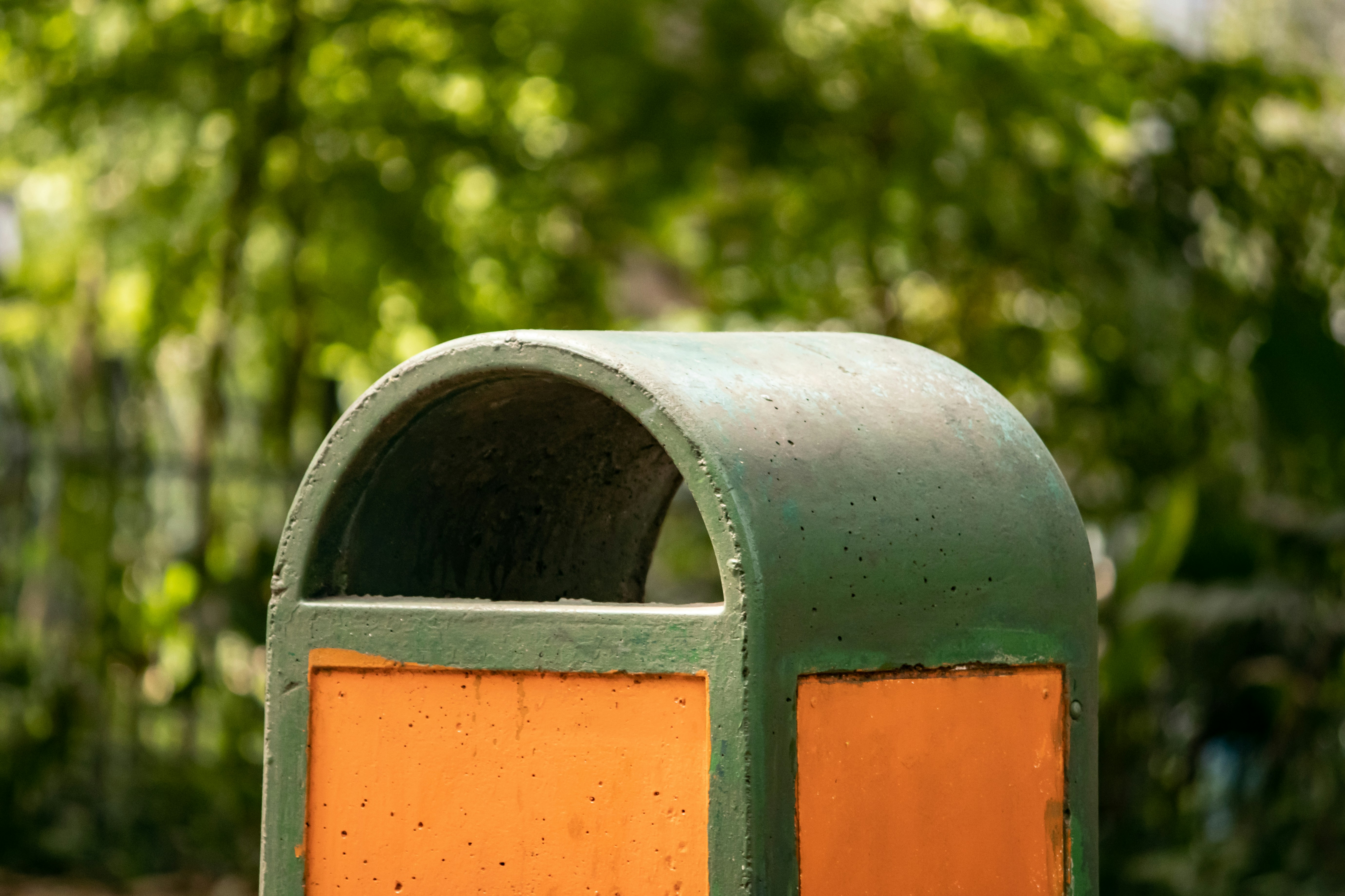The top of a public bin in a leafy park, with an arced green cover and orange sides.