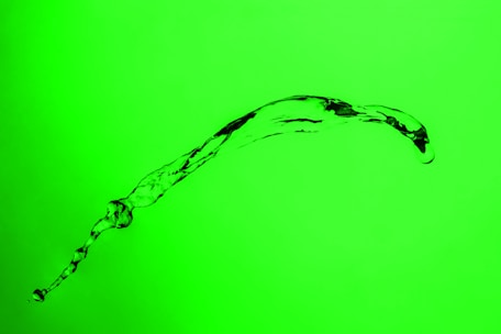 A stream of clear liquid is captured in mid-air against a bright green background. The liquid forms an arc, with smaller droplets and textured patterns visible in the flow.