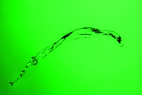 A stream of clear liquid is captured in mid-air against a bright green background. The liquid forms an arc, with smaller droplets and textured patterns visible in the flow.